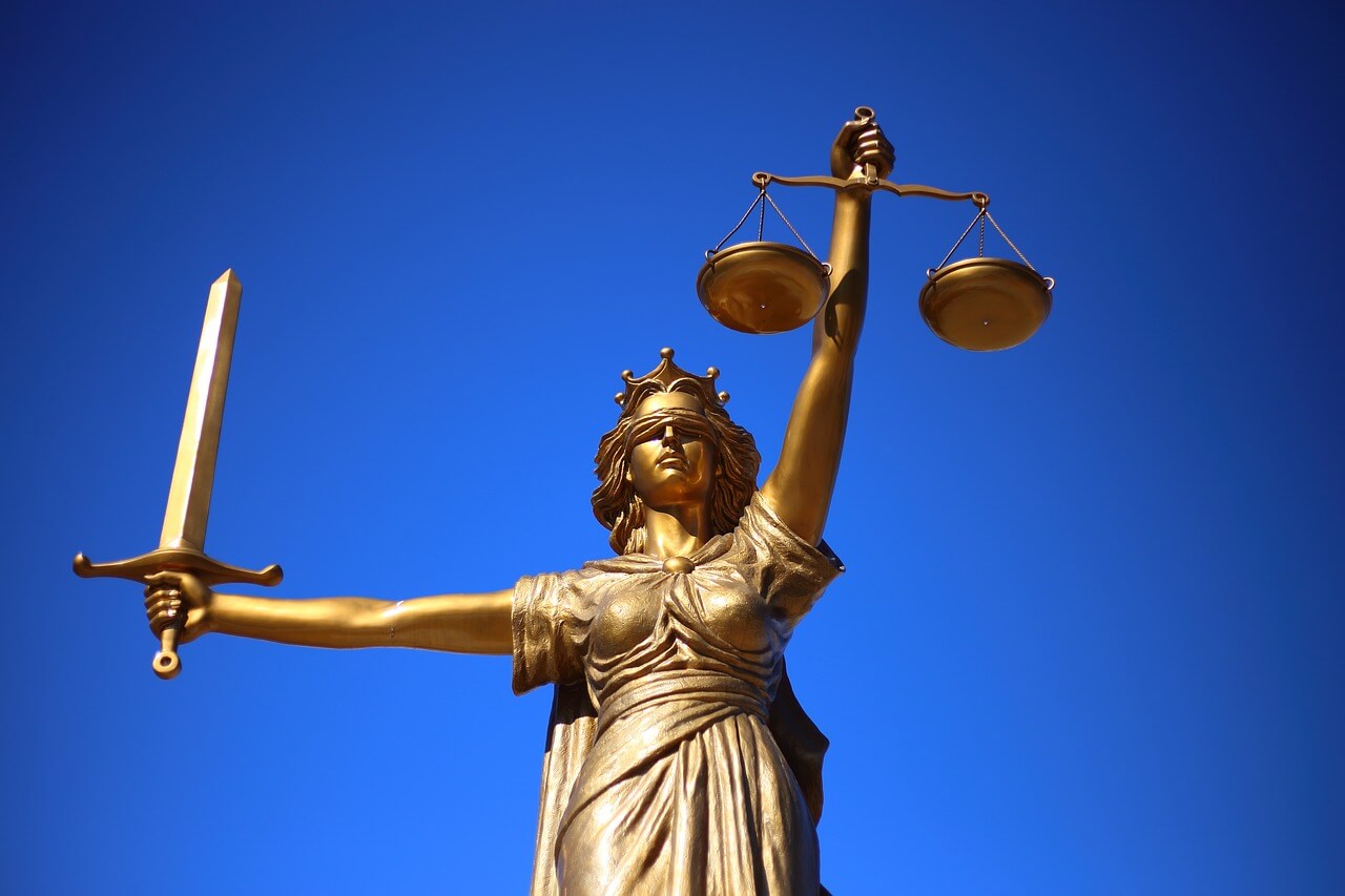 Bronzestatue von Justitia mit verbundenen Augen, die eine Waage und ein Schwert hält, vor einem klaren blauen Himmel.